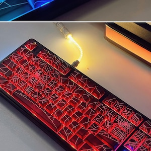 May include: A black mechanical keyboard with red keycaps and a spiderweb design. The keyboard is lit up with red and white lights.