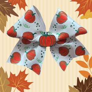 May include: A white hair bow featuring a repeating pattern of orange pumpkins and green vines. The bow is accented with small clear rhinestones and has a larger, central orange pumpkin embellishment at the knot of the bow.