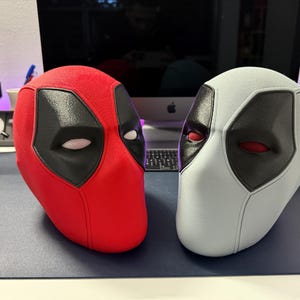Puede incluir: Dos máscaras de Deadpool, una roja y otra blanca, con agujeros negros para los ojos y ojos rojos. Las máscaras están hechas de un material texturizado y están sobre una superficie oscura.