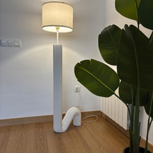 May include: A white floor lamp with a white fabric lampshade. The lamp has a unique, curved design and is standing on a wooden floor.