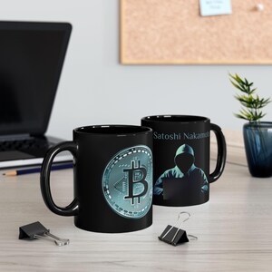 May include: Two black ceramic mugs with a Bitcoin logo and the text "Satoshi Nakamoto" on them. The mug on the left has a blue and green Bitcoin logo. The mug on the right has a silhouette of a person wearing a hooded sweatshirt and a laptop.