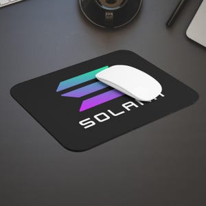 Może przedstawiać: Czarna prostokątna podkładka pod mysz z napisem "SOLANA" białymi literami. Podkładka pod mysz ma kolorowe logo z turkusowymi, fioletowymi i niebieskimi kształtami geometrycznymi. Na podkładce spoczywa biała mysz bezprzewodowa.
