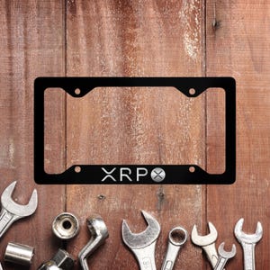 May include: Black metal license plate frame with the text "XRP" and a silver logo. The frame is on a wooden background.