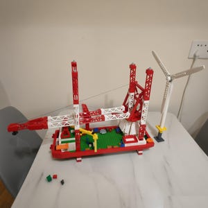 May include: A plastic building block construction set featuring a red and white offshore wind turbine installation vessel with a crane, a green deck, and a separate white three-bladed wind turbine model standing on a small gray base.