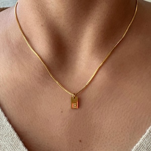 Op de afbeelding: Een goudkleurige ketting met een kleine rechthoekige hanger. De hanger heeft de letter "E" in een eenvoudig, elegant lettertype. De ketting wordt om de nek gedragen en de ketting heeft een delicaat design.