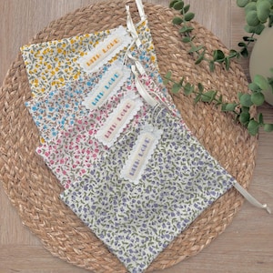 May include: Four floral fabric pouches in yellow, blue, pink, and purple, each with a white tag reading "WITH LOVE." The pouches are tied with white ribbons and are displayed on a woven mat, suitable for gifting or storage.