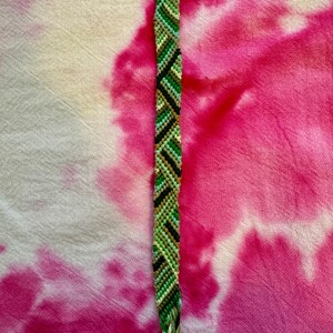 May include: A green, brown, and white friendship bracelet with a chevron pattern. The bracelet is lying on a pink and white tie-dye fabric.