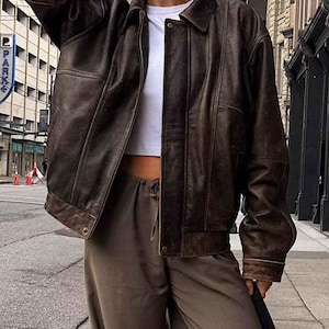 Peut inclure: Une veste bomber en cuir marron avec une fermeture éclair et un t-shirt blanc en dessous. La veste a un aspect vieilli et est portée sur un pantalon marron.