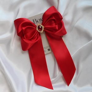 May include: A vibrant red satin bow with long streamers, centered with a gold-toned jewel. The bow is set against a white satin background, creating a striking contrast. The bow is a decorative accessory.