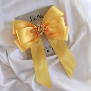 May include: A vibrant yellow satin bow with long tails, set against a grey card. The bow is adorned with a sparkling, oval-shaped jewel. The card displays the word "BOW" and the word "INSPIRATION". The bow is on a white, silky background.