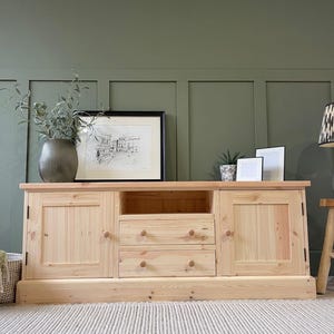 May include: A natural pine entertainment unit with two doors, two drawers, and an open shelf. The unit is set against a green wall. A framed picture and decorative items are displayed on top. The unit is approximately 150 cm wide.