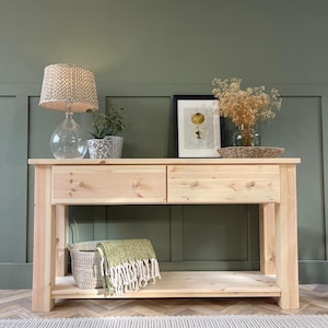 May include: A light wood console table with two drawers and a lower shelf. The table is in front of a sage green wall and has a white woven basket and a green throw blanket on the shelf. There is a lamp with a patterned shade and a potted plant on the table.