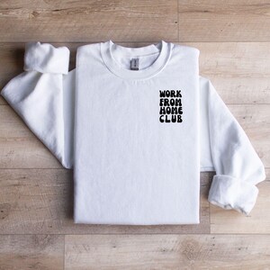 K&ouml;nnte beinhalten: Wei&szlig;er Rundhals-Pullover mit dem Aufdruck "WORK FROM HOME CLUB" auf der linken Brust.