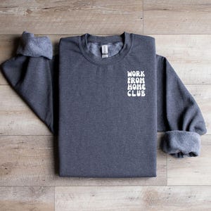 K&ouml;nnte beinhalten: Ein dunkelgrauer Pullover mit dem Text "WORK FROM HOME CLUB" in Wei&szlig; auf der Vorderseite.
