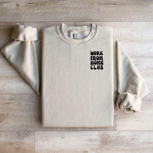 K&ouml;nnte beinhalten: Ein hellbeiger Pullover mit dem Aufdruck "WORK FROM HOME CLUB" in Schwarz auf der Vorderseite.