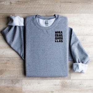 K&ouml;nnte beinhalten: Grauer Pullover mit dem Aufdruck "WORK FROM HOME CLUB" auf der Vorderseite.