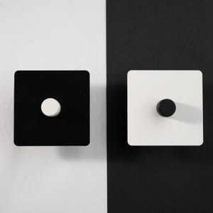 Könnte beinhalten: Zwei quadratische, minimalistische Schrankknöpfe. Einer ist schwarz mit einem weißen runden Knopf, der andere ist weiß mit einem schwarzen runden Knopf. Der Hintergrund ist geteilt, mit Weiß links und Schwarz rechts.