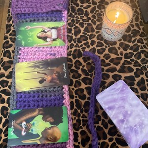 May include: Three tarot cards with purple, pink, and gray crocheted fabric surrounding them. The cards are "Ten of Pentacles", "Four of Wands", and "Two of Cups". A lit candle and a purple and white box are on a brown and black leopard print fabric.