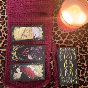 May include: Three tarot cards, Swords, Cups, and Wands, laid out on a burgundy crocheted bag. A deck of tarot cards is to the right of the bag on a leopard print surface. A lit candle is in the upper right corner of the image.