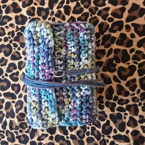 May include: A handmade crochet journal cover in shades of blue, purple, and green, secured with a gray cord and metal ring closure. The cover is displayed on a brown and black leopard print background.