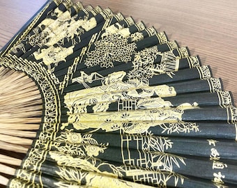 May include: A black and gold folding fan with intricate Asian-inspired illustrations. The fan features detailed gold designs on a black background, with a bamboo handle. The fan is partially open, displaying the detailed artwork.