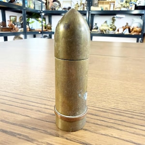 May include: A vintage brass bullet-shaped container with a pointed top and a cylindrical body. The container has a two-part design, with a visible seam. A copper-coloured ring encircles the base. The item is displayed on a wooden surface.