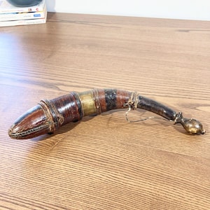 Op de afbeelding: Een handgemaakte, gebogen dolk met een met leer omwikkelde handgreep en een decoratieve metalen pommel. De handgreep heeft banden van bruine, zwarte en goudkleurige materialen. Het lemmet is bedekt met een bruine leren schede.