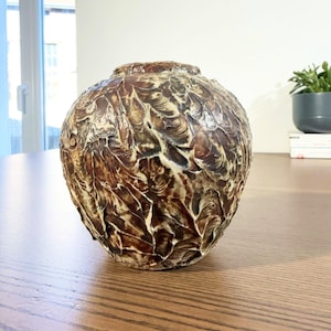 Puede incluir: Un jarrón redondo decorativo con una superficie texturizada. El jarrón tiene una combinación de colores marrón y crema, con un patrón de formas superpuestas en forma de hoja. El jarrón está sobre una mesa de madera.