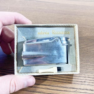 May include: A vintage silver Adonis Ronson lighter in its original box. The lighter has a rectangular shape with a textured surface and a hinged top. The box is light green with the brand name printed in gold.
