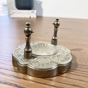 Può includere: Un porta orologio in metallo argentato con due pilastri decorativi e una base a forma di conchiglia. La base presenta un intricato disegno floreale.