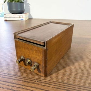 Puede incluir: Una caja rectangular de madera con tapa con bisagras y tres perillas de latón en la parte delantera. La caja es de madera marrón claro con una tapa marrón más oscura. Las perillas son de color dorado brillante. Es probable que la caja sea una radio antigua o un dispositivo electrónico.