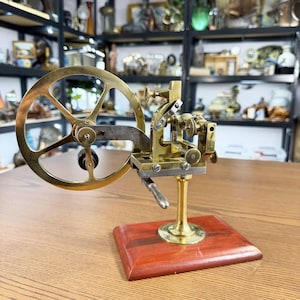Può includere: Un vecchio tornio da orologiaio in ottone e acciaio, con una grande ruota a raggi e ingranaggi intricati. Il tornio è montato su una base di legno marrone scuro. Lo sfondo mostra una collezione di oggetti antichi. Il tornio è di colore dorato.