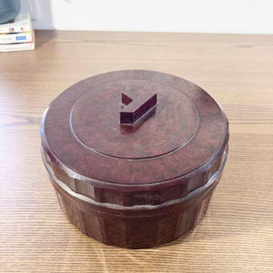 May include: A circular, dark brown box with a lid and a Y-shaped handle. The box has a textured surface and a geometric design around the base. The lid is circular and fits snugly on top of the box.
