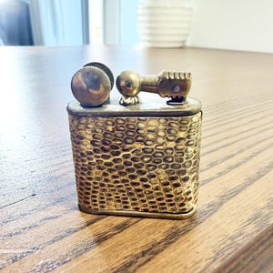 May include: A vintage brass lighter with a textured, scale-like pattern on the body. The lighter has a round, textured knob and a rectangular top with a lever. The metal has a warm, antique gold colour, suggesting age and use.