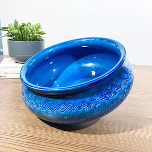 Op de afbeelding: Een levendige blauwe keramische kom met een glanzende afwerking. De kom heeft een unieke, asymmetrische vorm en een gestructureerd, geometrisch patroon rond de basis. De kom wordt waarschijnlijk gebruikt voor decoratieve doeleinden of als plantenbak.