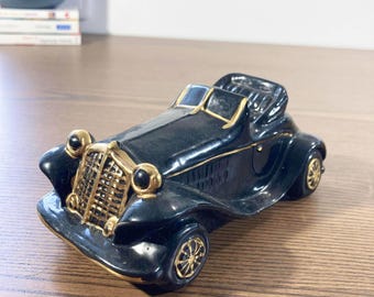 May include: A black vintage car ornament with gold detailing. The classic car features a detailed front grille, headlights, and wheels. The ornament is made of a glossy material and is placed on a wooden surface.