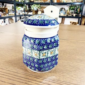Könnte beinhalten: Ein Keramiktopf mit Deckel, verziert mit einem blau-grünen Blumenmuster. Der Topf hat einen weißen Rand und einen passenden Deckel mit dunkelblauem Rand. Das Design umfasst kleine gelbe und grüne Blumenakzente. Der Topf steht auf einer Holzoberfläche.