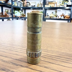 Puede incluir: Un telescopio antiguo de latón con un diseño cilíndrico. El telescopio tiene un aspecto deslustrado y envejecido, con grabados detallados alrededor de la sección media. La base presenta un diseño estriado. El telescopio está de pie sobre una superficie de madera.