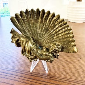 Peut inclure: Un objet décoratif en métal doré en forme de grand éventail ouvert. L'éventail présente une sculpture en relief d'une silhouette drapée dans un tissu fluide, positionnée horizontalement sur la partie inférieure de la structure plissée.
