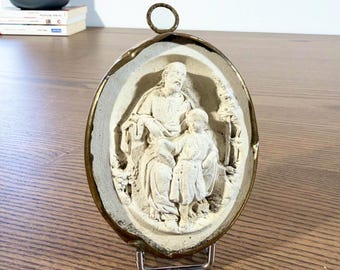 May include: Oval relief sculpture in off-white, featuring a seated figure and a child, framed by a gold-coloured metal edge. The sculpture is mounted on a metal stand, suitable for display.