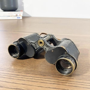 May include: Antique black binoculars with brass detailing and a leather strap. The binoculars are on a wooden surface. The lenses are black and the body shows signs of wear and tear.