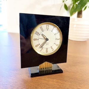 Könnte beinhalten: Eine quadratische schwarze Tischuhr mit einem runden cremefarbenen Zifferblatt, goldenen Ziffern und goldenen Zeigern. Die Uhr verfügt über einen goldenen metallischen Akzent am Sockel und ist in einen glänzenden schwarzen Rahmen mit passendem Ständer eingefasst.