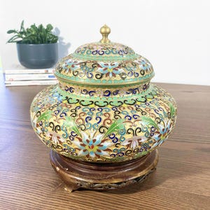 May include: A decorative, lidded jar with intricate floral and swirl patterns in gold, green, blue, and red. The jar sits on a round, wooden stand. The lid is topped with a small, gold finial. The overall design is ornate and detailed.