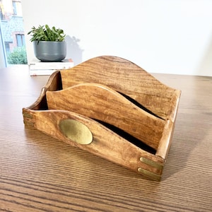 May include: A wooden desk organiser with three compartments. The organiser is made of light brown wood with a wavy top and brass accents. A small brass oval is on the front. The organiser is on a wooden table.