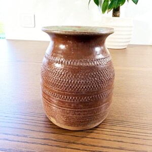 Peut inclure: Un vase en céramique marron avec un bord évasé et un corps arrondi. La surface extérieure présente des bandes horizontales de motifs texturés et imprimés, incluant des croisillons et des lignes en zigzag, avec un vernis brillant.
