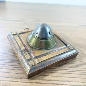May include: Vintage inkwell featuring a square wooden base and a metal ink pot. The ink pot has a conical top and a brass base with the number "1006" engraved. The wooden base has a decorative border and a small metal loop for hanging.
