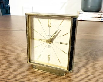 May include: A vintage rectangular clock with a cream-colored face and gold-tone hands and accents. The clock face features black numerals and markings, with the number 12 prominently displayed. The clock is set on a gold-tone base and has black side panels.