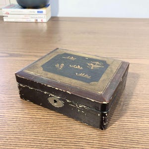 Peut inclure: Une petite boîte en bois avec un couvercle doré et des détails peints en noir. Le couvercle présente un motif de feuilles stylisées et un oiseau. La boîte a un trou de serrure sur le devant.