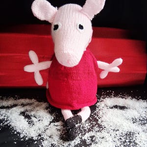 May include: A knitted pink pig doll wearing a red dress and black shoes. The pig has large ears, black eyes, and outstretched arms. The doll is positioned on a surface with white flakes, with a red backdrop.