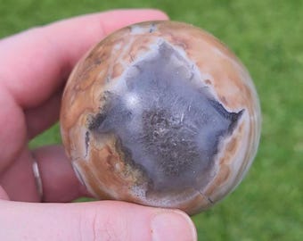 May include: A polished stone sphere with a mix of tan, brown, and gray colors. The sphere has a prominent gray and white pattern in the center, resembling a star or flower. The stone is held in a hand against a blurred green background.